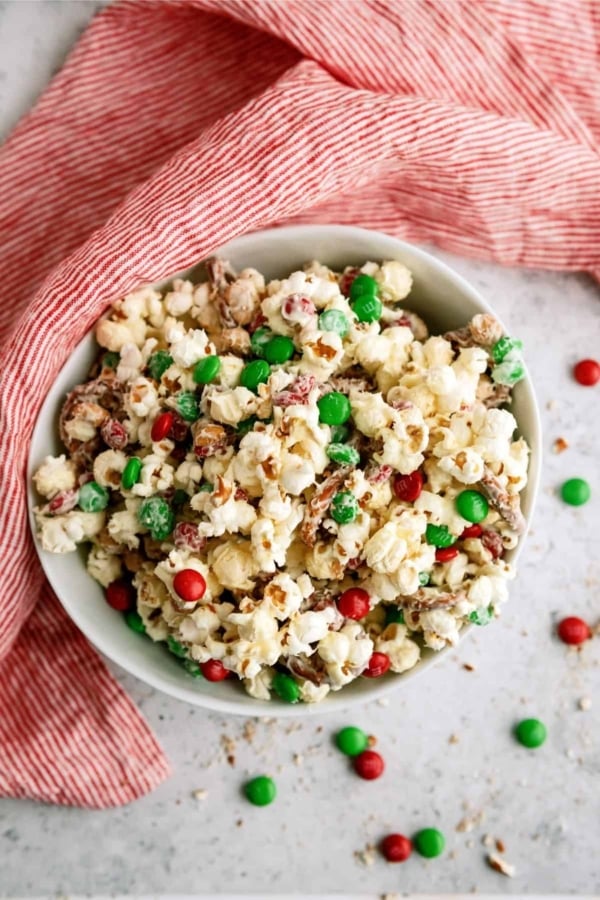 A bowl of popcorn mixed with red and green candy-coated chocolates, placed on a red-striped cloth.