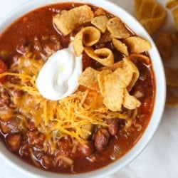 A bowl of chili topped with shredded cheese, sour cream, and corn chips, with pieces of cornbread on the side.