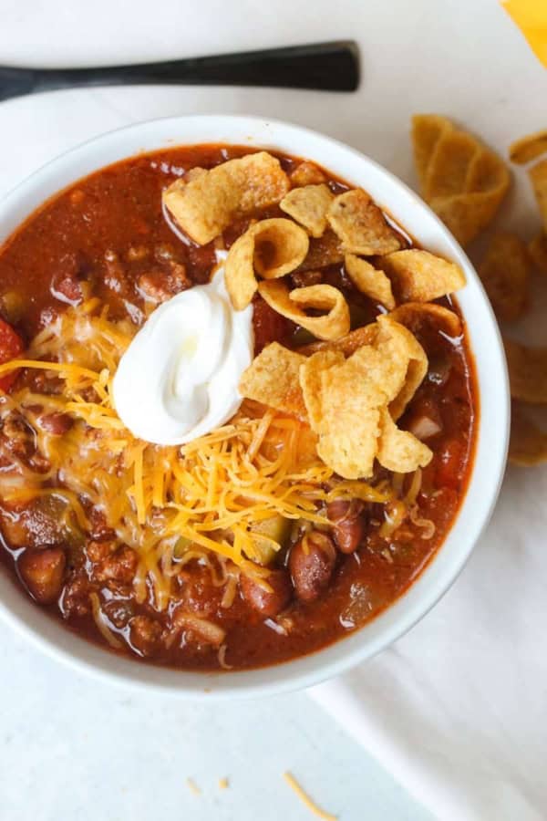 A bowl of chili topped with shredded cheese, sour cream, and corn chips, with pieces of cornbread on the side.