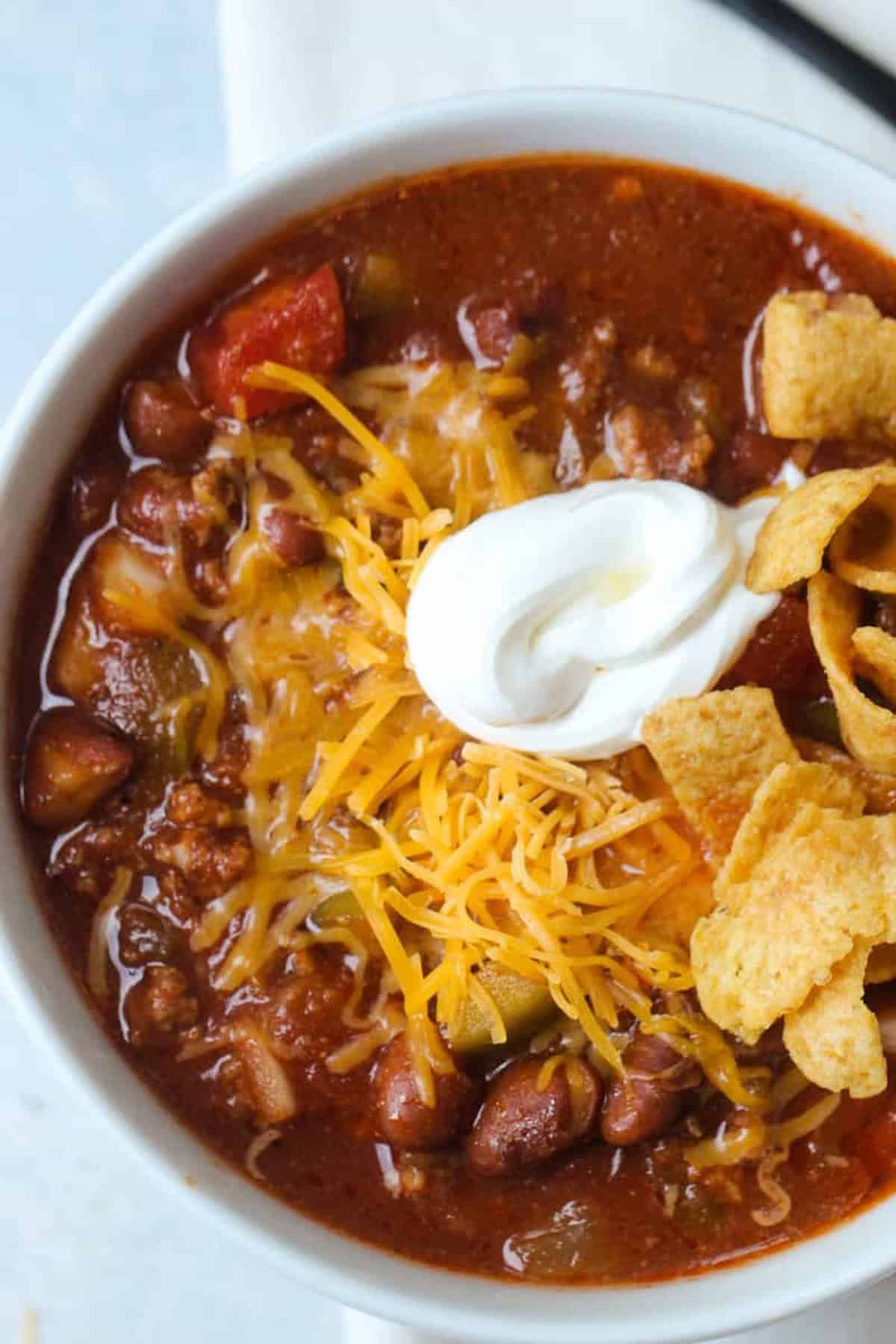 A bowl of chili topped with shredded cheese, a dollop of sour cream, and corn chips. Beans and ground meat are visible in the chili.