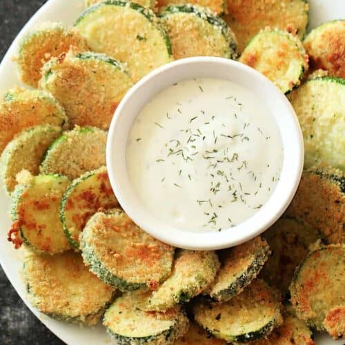 A plate of breaded and fried zucchini slices surrounding a small bowl of creamy dipping sauce garnished with herbs.