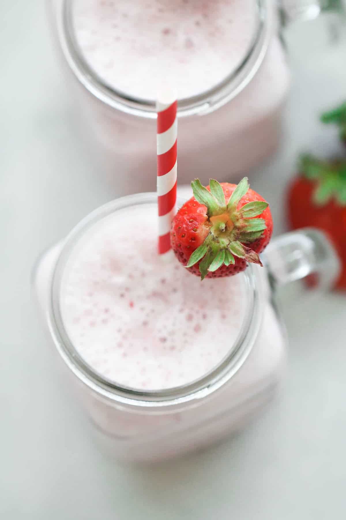 A glass mug filled with a frothy pink strawberry smoothie, topped with a red and white striped straw and a whole strawberry on the rim.