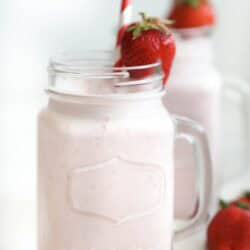 A glass jar filled with a light pink strawberry milkshake, garnished with a whole strawberry and a red-and-white striped straw; another similar jar in the background.