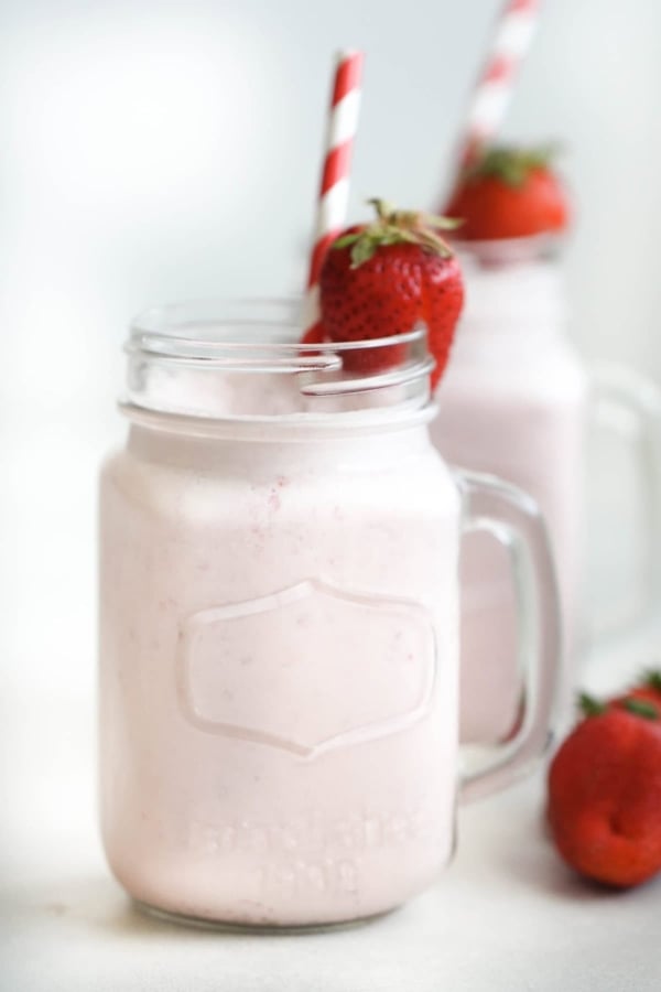A glass jar filled with a light pink strawberry milkshake, garnished with a whole strawberry and a red-and-white striped straw; another similar jar in the background.
