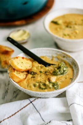 A ceramic bowl filled with creamy soup, featuring pieces of broccoli and bread slices, with a black spoon resting inside. Another similar bowl and a small dish of butter in the background.