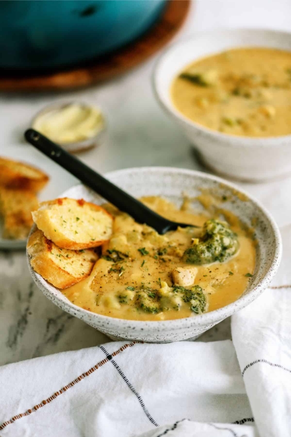A ceramic bowl filled with creamy soup, featuring pieces of broccoli and bread slices, with a black spoon resting inside. Another similar bowl and a small dish of butter in the background.