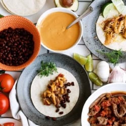 A variety of taco ingredients including tortillas, black beans, cheese sauce, chopped tomatoes, beef, shredded cheese, avocado, sour cream, and lime wedges arranged on a table.