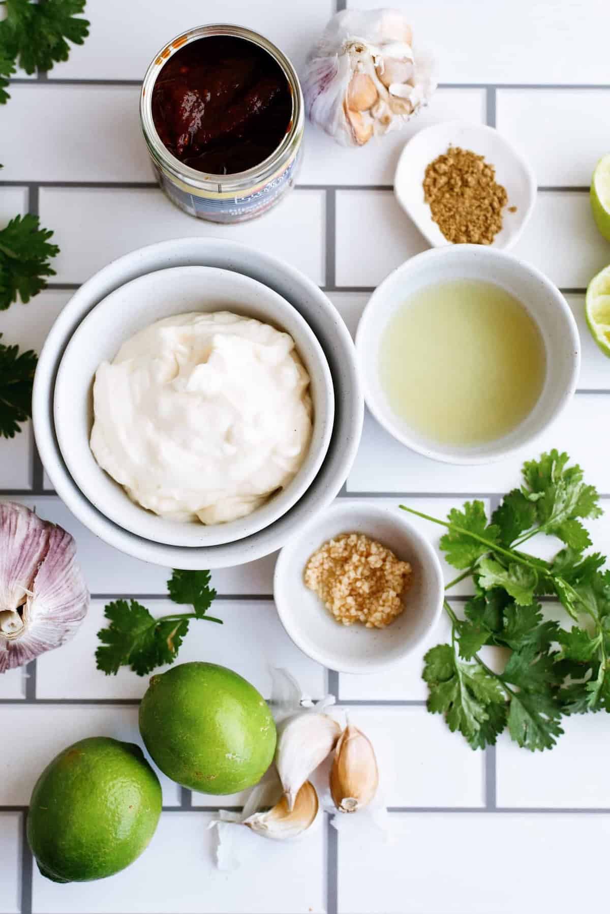 Ingredients arranged on a tiled surface, including mayonnaise, limes, garlic, cilantro, ground spices, chipotle peppers in adobo sauce, and a small bowl of lime juice.