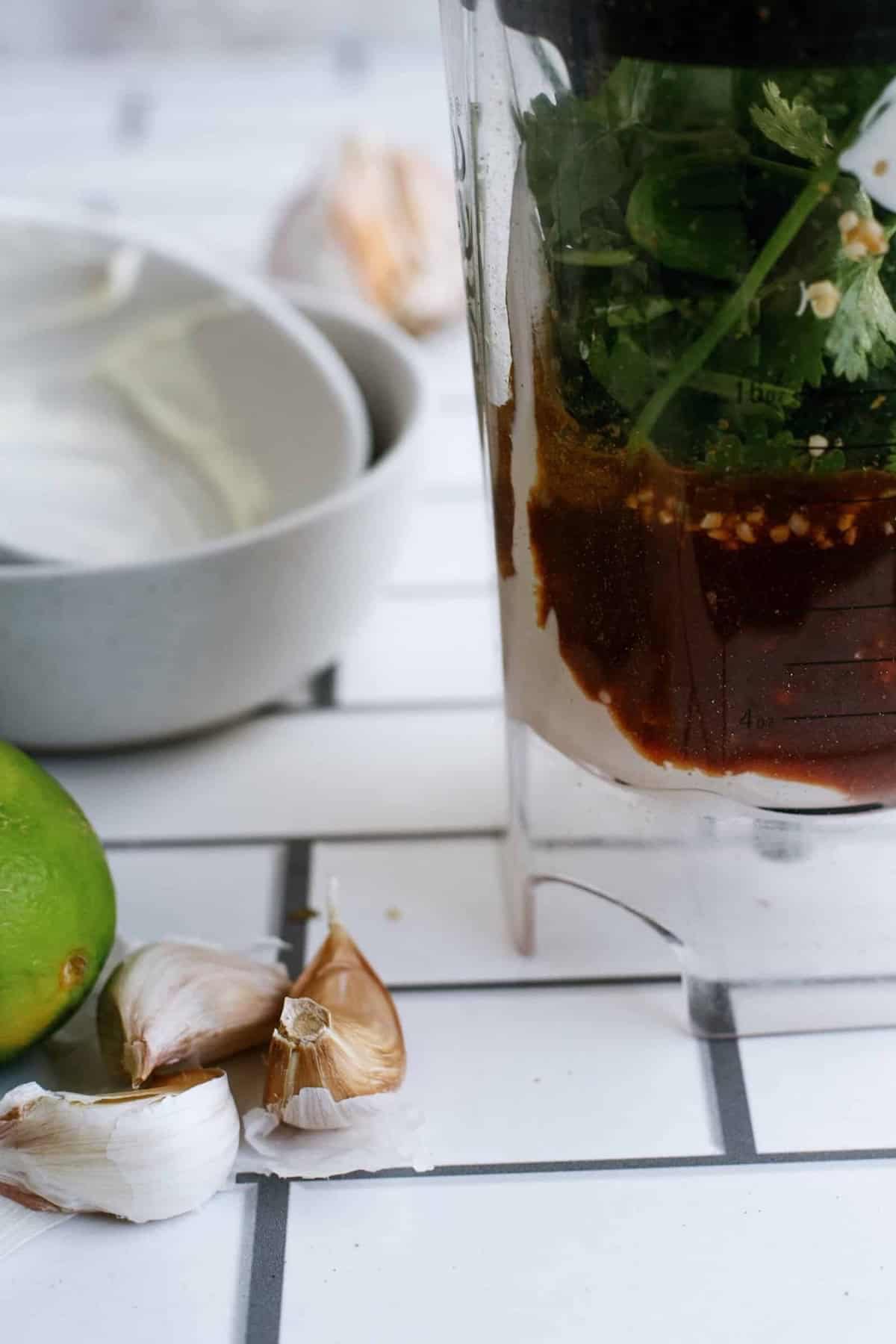 A blender container filled with herbs and sauces sits on a white tiled counter next to garlic cloves, a lime, and stacked white bowls.