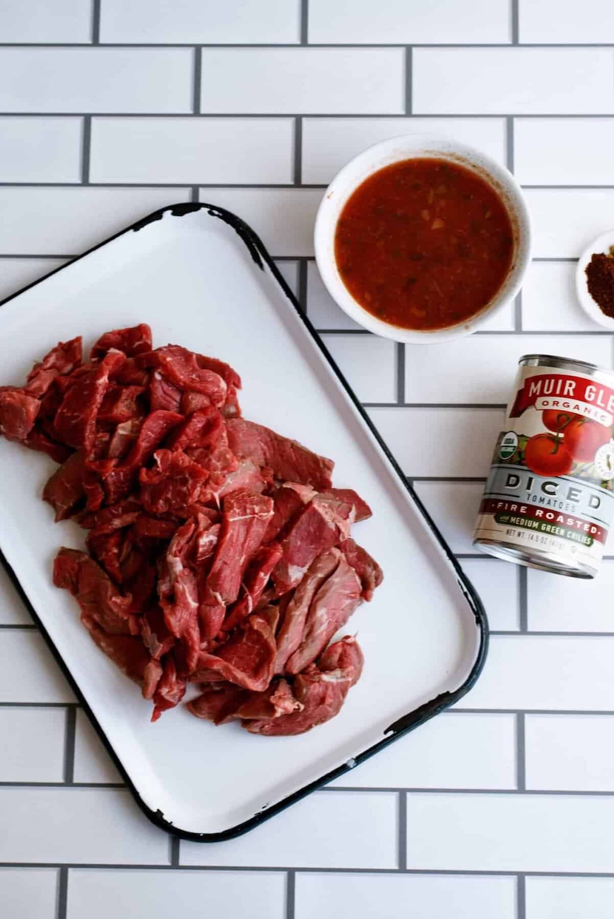 Sliced raw beef on a white tray, a can of diced tomatoes, a bowl of tomato sauce, and a small bowl of spices on a white tile surface.