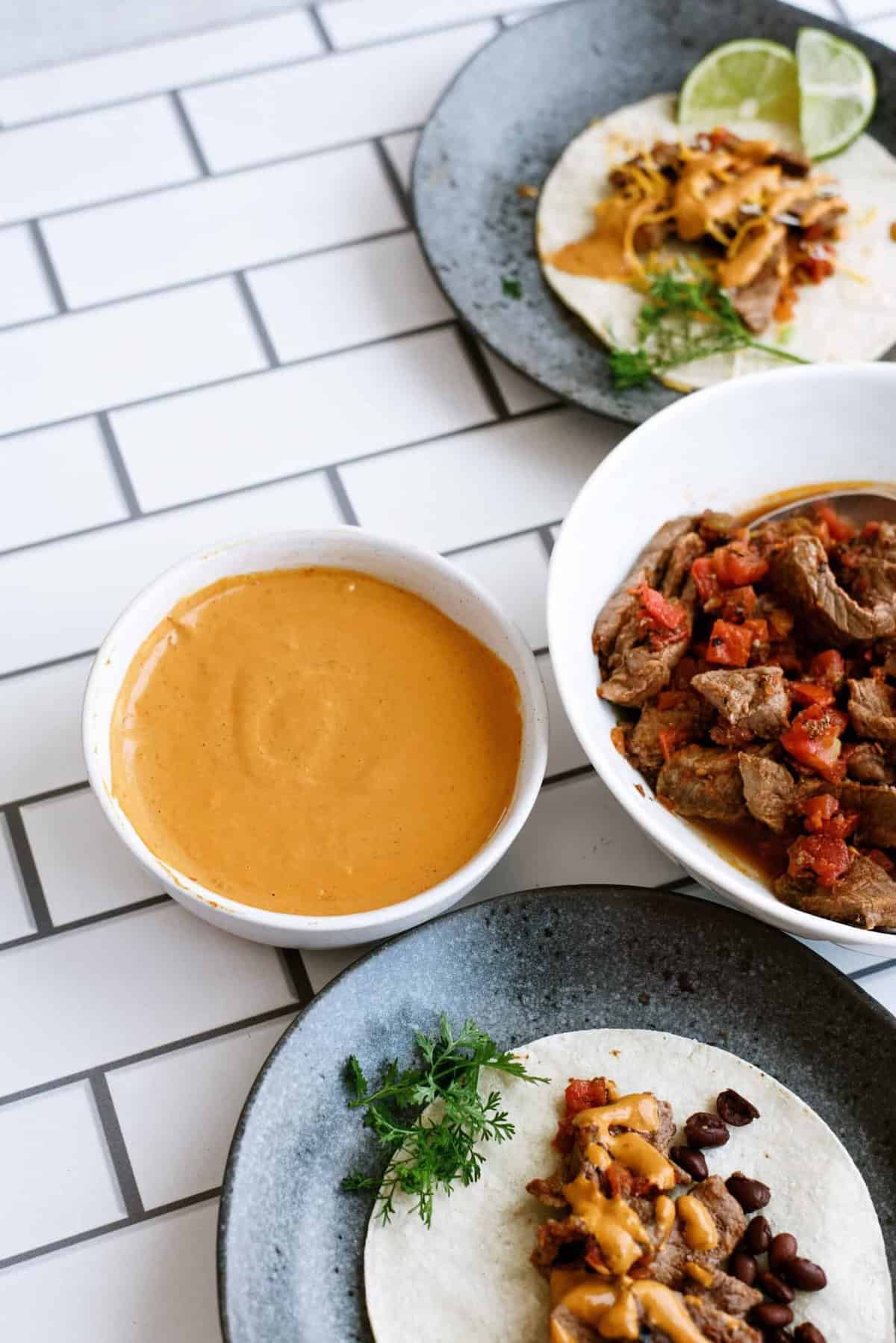 A bowl of cheese sauce sits next to plates with tortillas topped with beans, melted cheese, beef, and diced tomatoes on a white tiled surface.