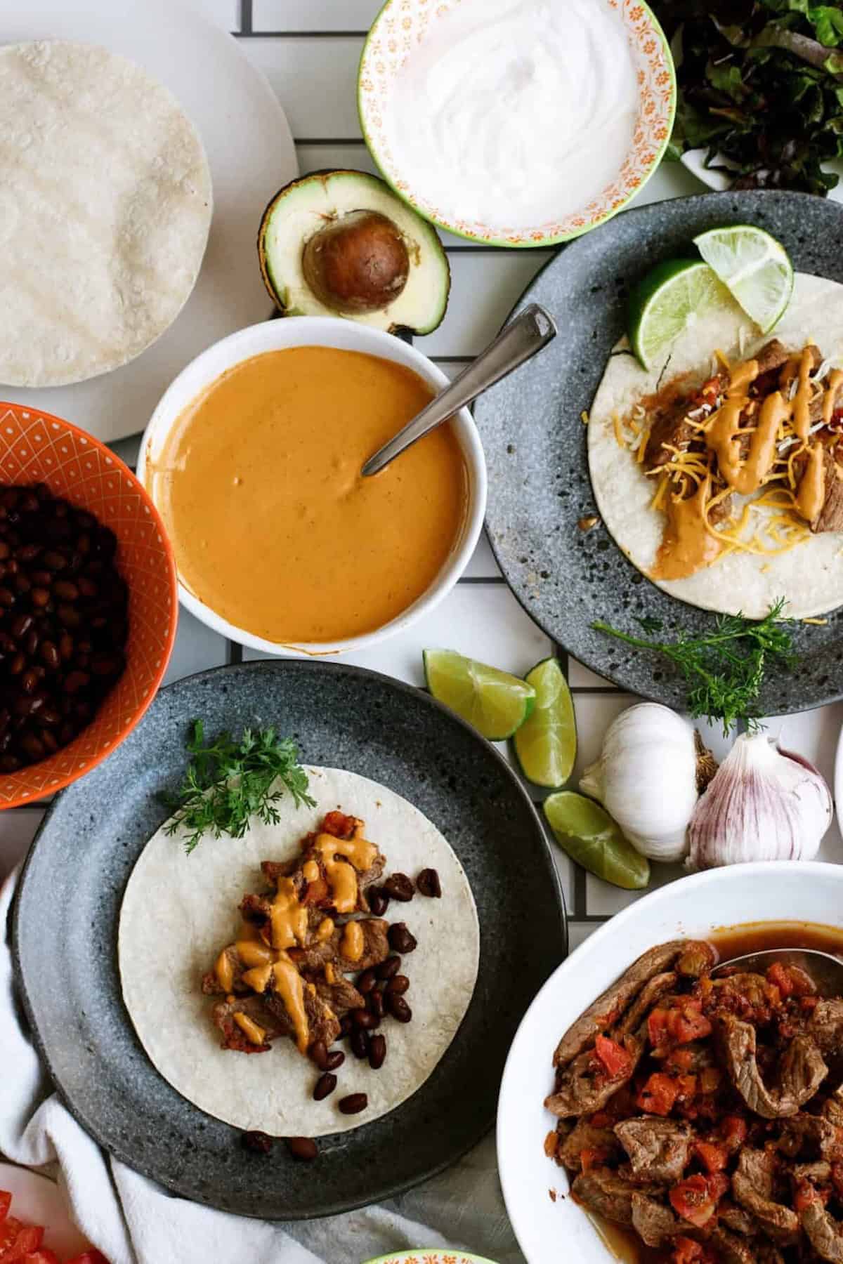 Overhead view of tortillas with beef, beans, and cheese sauce, surrounded by bowls of black beans, cheese sauce, sour cream, avocado, lime wedges, and cooked beef with tomatoes.