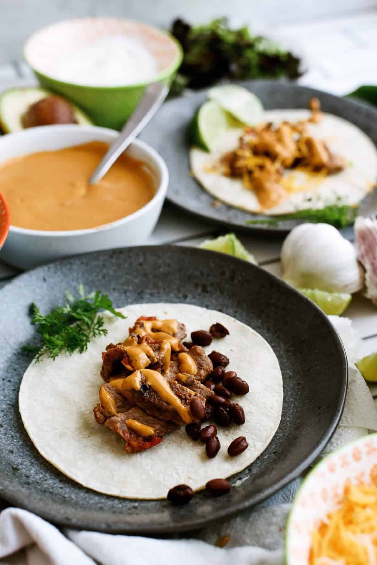 A soft taco shell on a plate topped with shredded meat, black beans, and cheese, with various bowls of sauces and ingredients in the background.