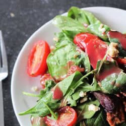 A plate of fresh spinach salad with grape tomatoes, crumbled cheese, and vinaigrette dressing on a dark countertop.