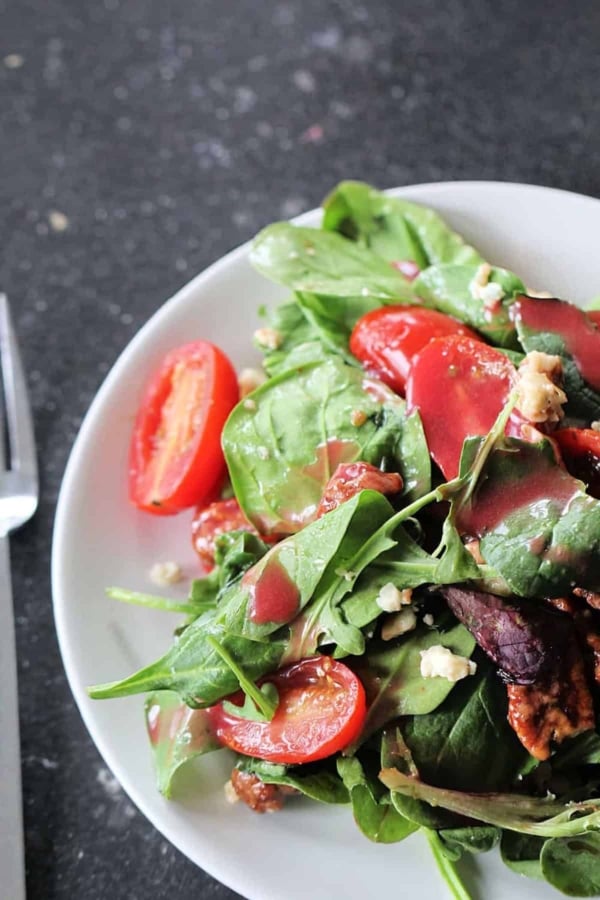 A plate of fresh spinach salad with grape tomatoes, crumbled cheese, and vinaigrette dressing on a dark countertop.
