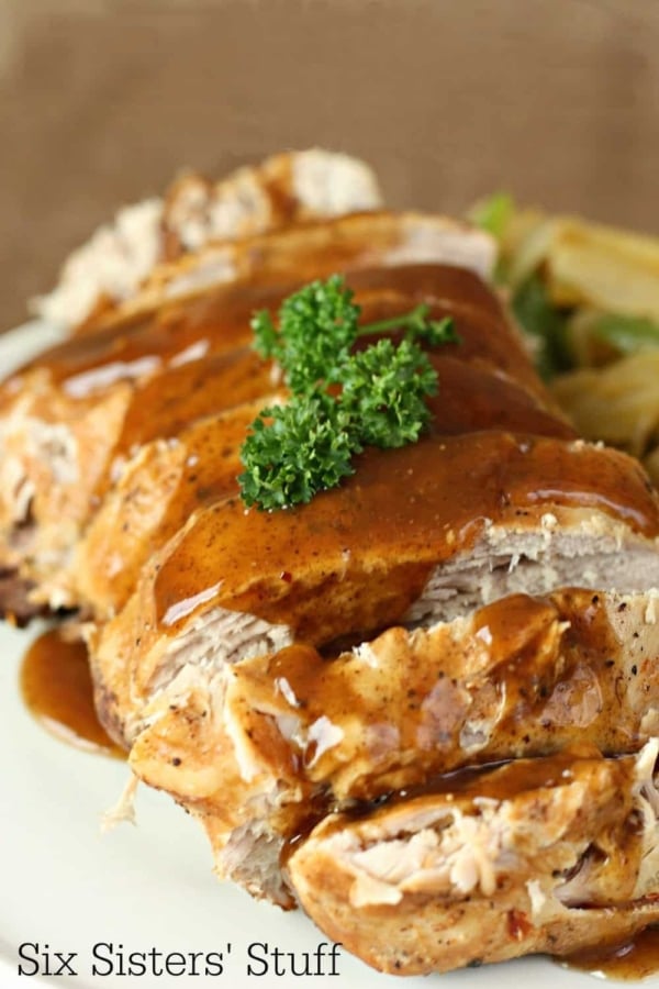 Close-up of sliced roast pork topped with brown gravy and garnished with parsley, served with a side of vegetables.