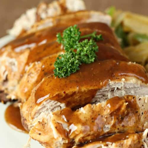 Close-up of sliced roast pork topped with brown gravy and garnished with parsley, served with a side of vegetables.