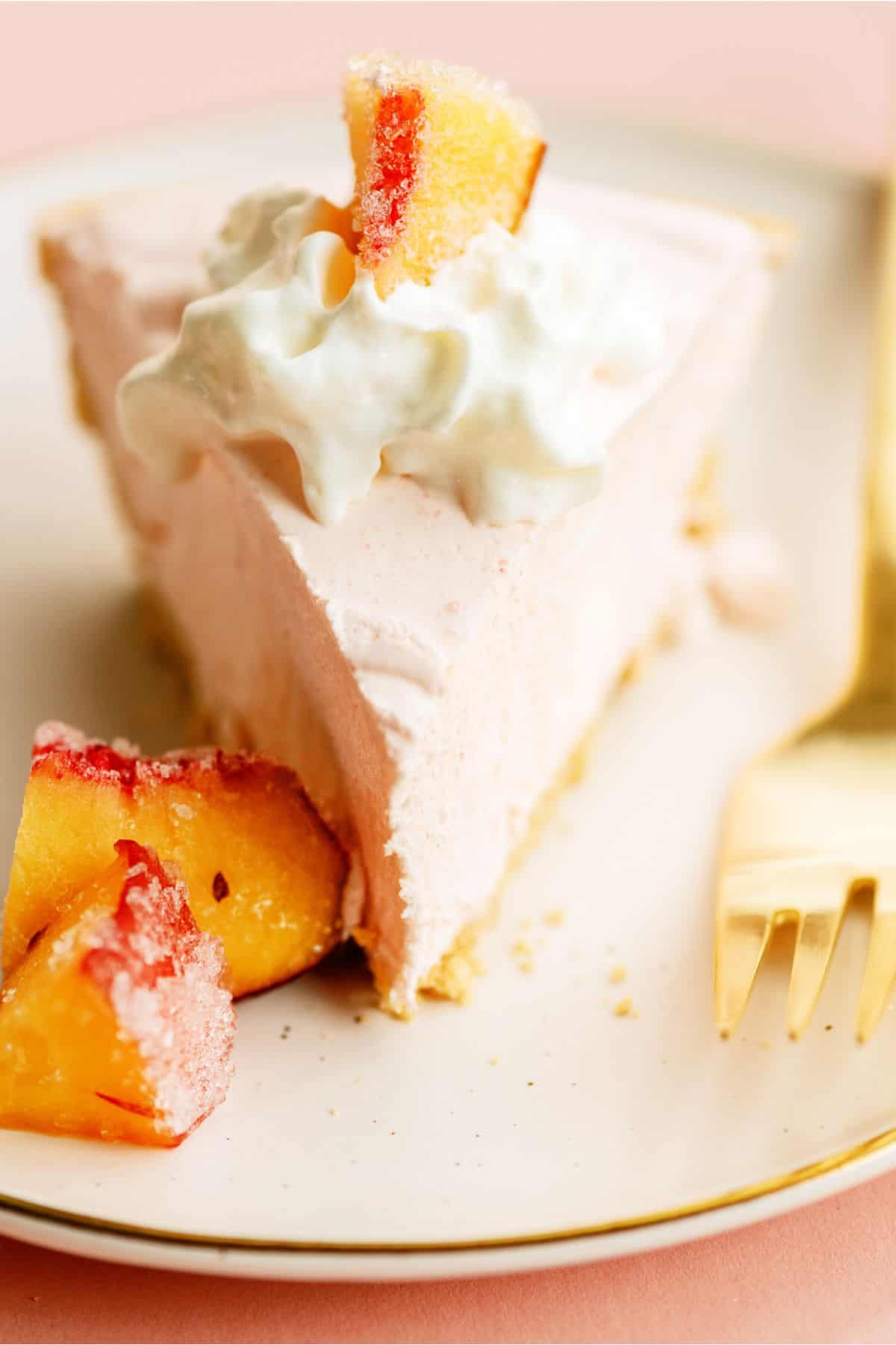 A slice of peach pie topped with whipped cream and sugared peach slices, served on a white plate with a gold fork.