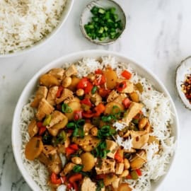 A bowl of rice topped with stir-fried chicken, red bell peppers, and green onions. Nearby are a bowl of rice, a small dish of chopped green onions, and a dish of red pepper flakes on a marble surface.