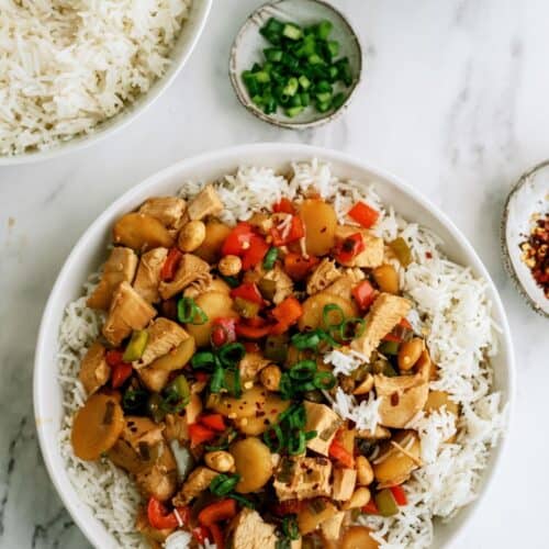 A bowl of rice topped with stir-fried chicken, red bell peppers, and green onions. Nearby are a bowl of rice, a small dish of chopped green onions, and a dish of red pepper flakes on a marble surface.