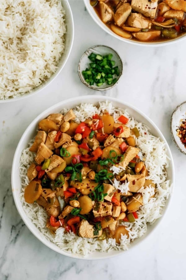 A bowl of rice topped with stir-fried chicken, red bell peppers, and green onions. Nearby are a bowl of rice, a small dish of chopped green onions, and a dish of red pepper flakes on a marble surface.