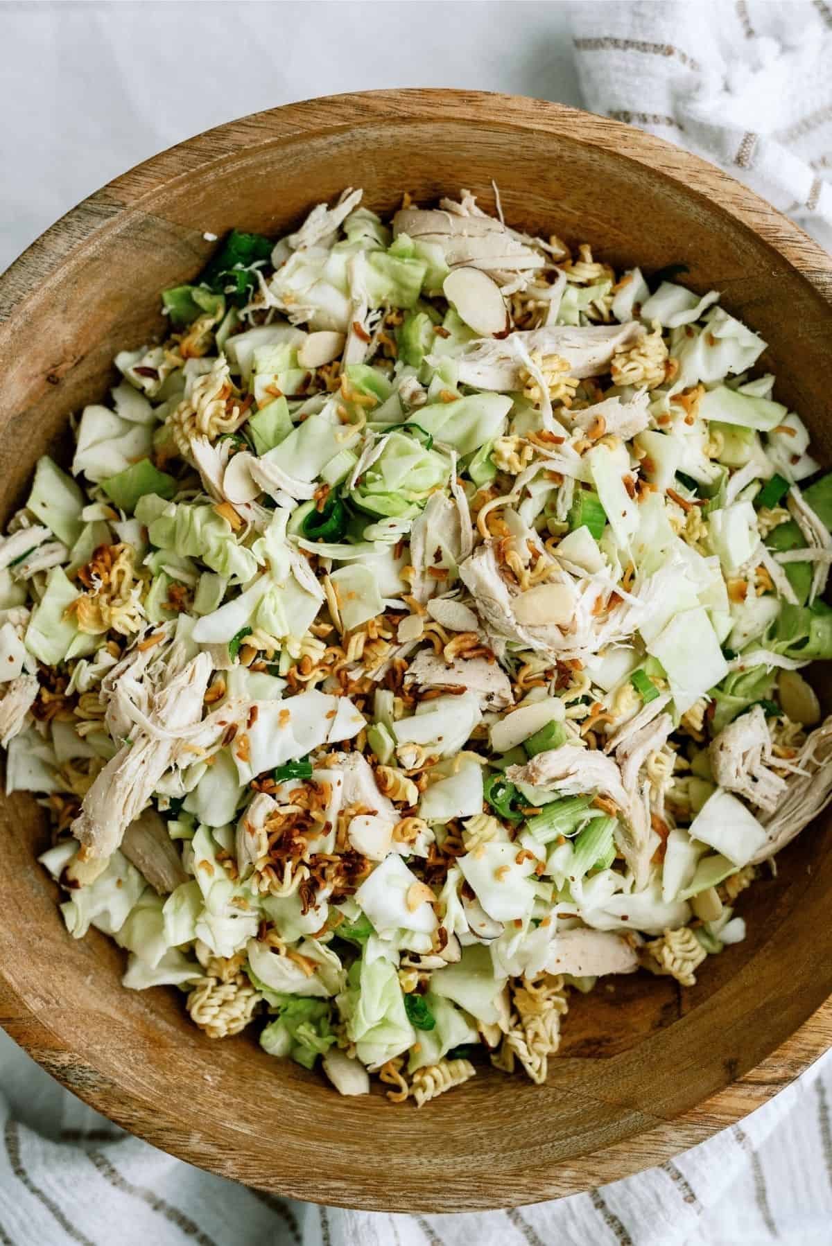 A wooden bowl filled with a salad of chopped cabbage, shredded chicken, sliced almonds, crushed instant noodles, and green onions.