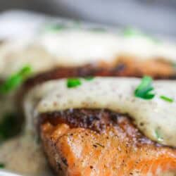 Close-up of a piece of seared salmon topped with a creamy white sauce and garnished with chopped green herbs on a white plate.