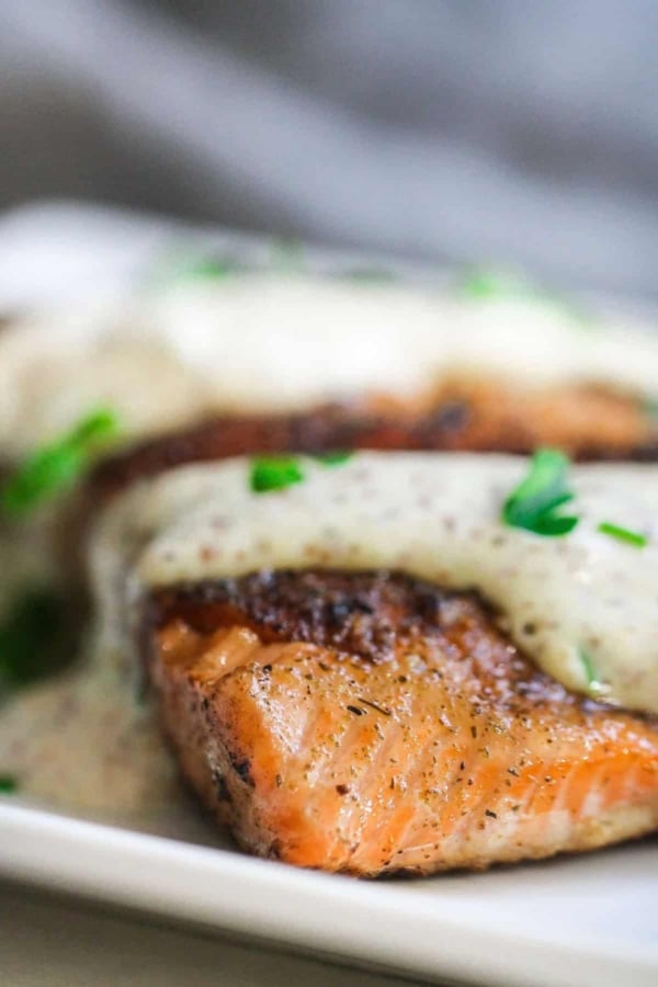 Close-up of a piece of seared salmon topped with a creamy white sauce and garnished with chopped green herbs on a white plate.