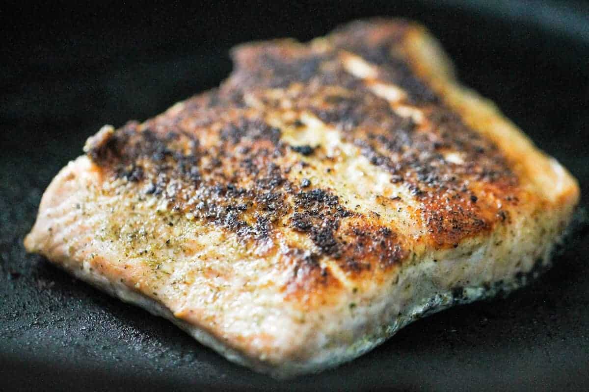 A close-up of a seasoned, pan-seared salmon fillet cooking in a skillet.