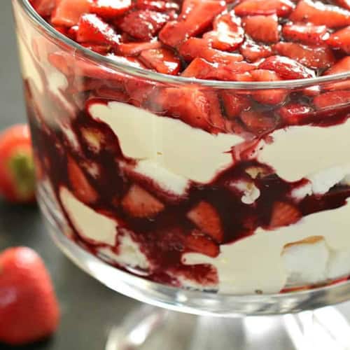 A glass trifle dish filled with layers of cream, strawberry sauce, and fresh strawberries, with whole strawberries placed around the base.