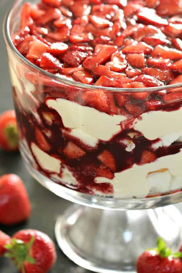 A glass trifle dish filled with layers of cream, strawberry sauce, and fresh strawberries, with whole strawberries placed around the base.