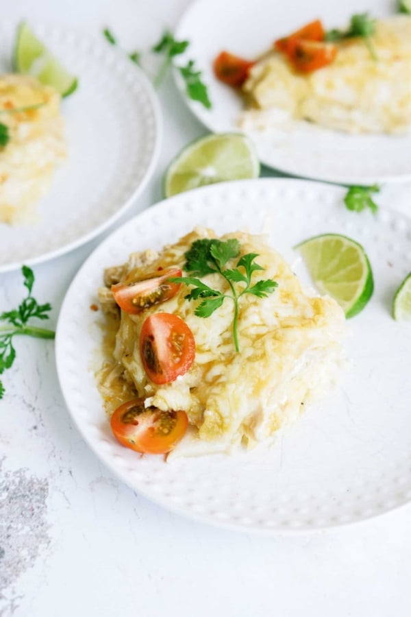 A slice of creamy casserole topped with cherry tomatoes and herbs on a white plate, garnished with lime wedges, with another plate of food in the background.