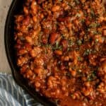 A close-up of baked beans with ground meat and herbs in a black skillet, placed on a light wood surface next to a blue and white striped cloth.