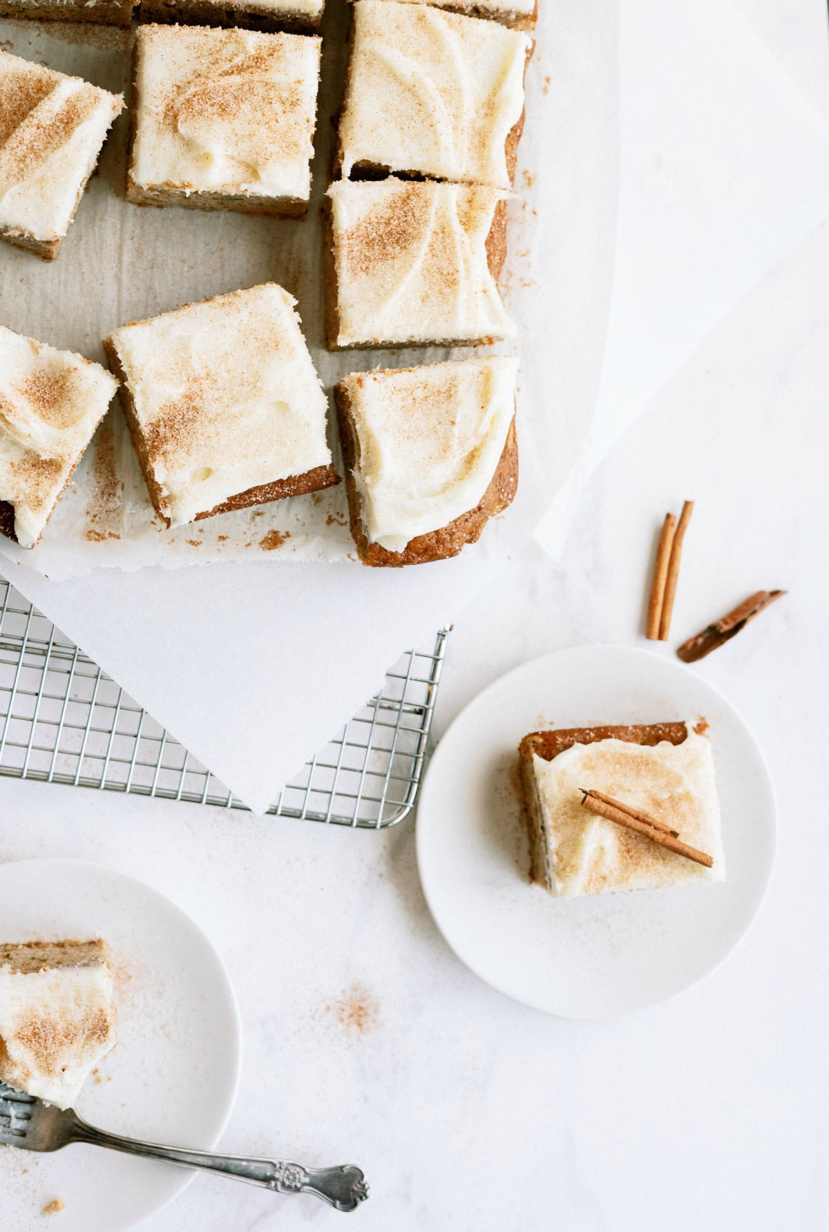 Overhead view of frosted cake squares on a cooling rack with one piece on a white plate garnished with a cinnamon stick.