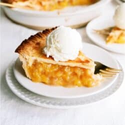 A slice of Fresh Classic Peach Pie on a plate topped with ice cream, the remaining pie in the background.