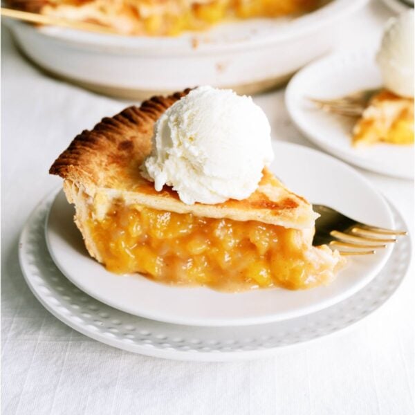 A slice of Fresh Classic Peach Pie on a plate topped with ice cream, the remaining pie in the background.