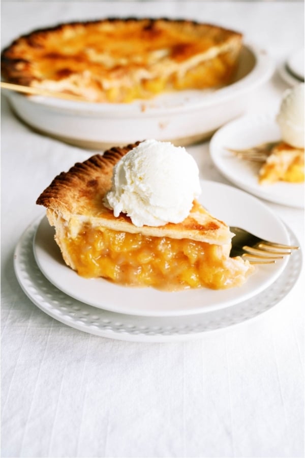 A slice of Fresh Classic Peach Pie on a plate topped with ice cream, the remaining pie in the background.
