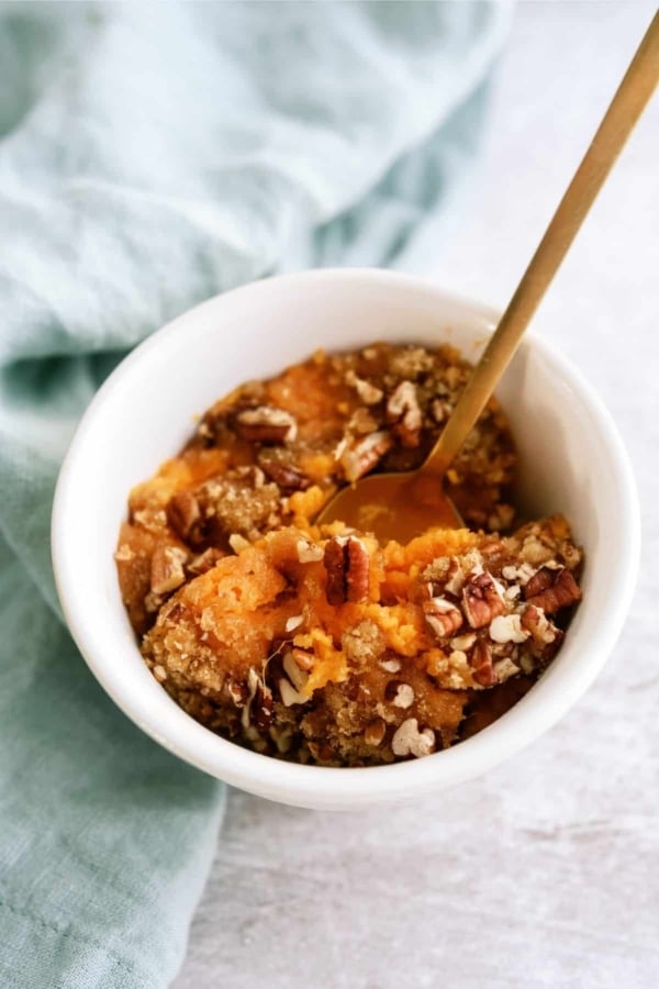 A white bowl filled with sweet potato casserole topped with pecans and a spoon in it on a cloth surface.