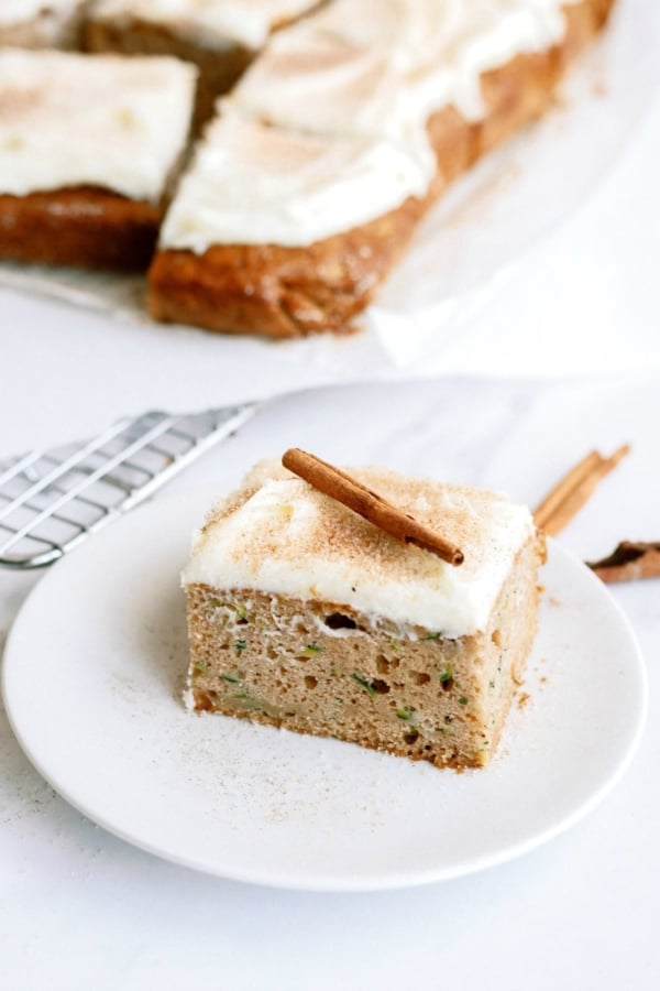 A slice of cake topped with frosting and a cinnamon stick is placed on a white plate, with the rest of the cake in the background.