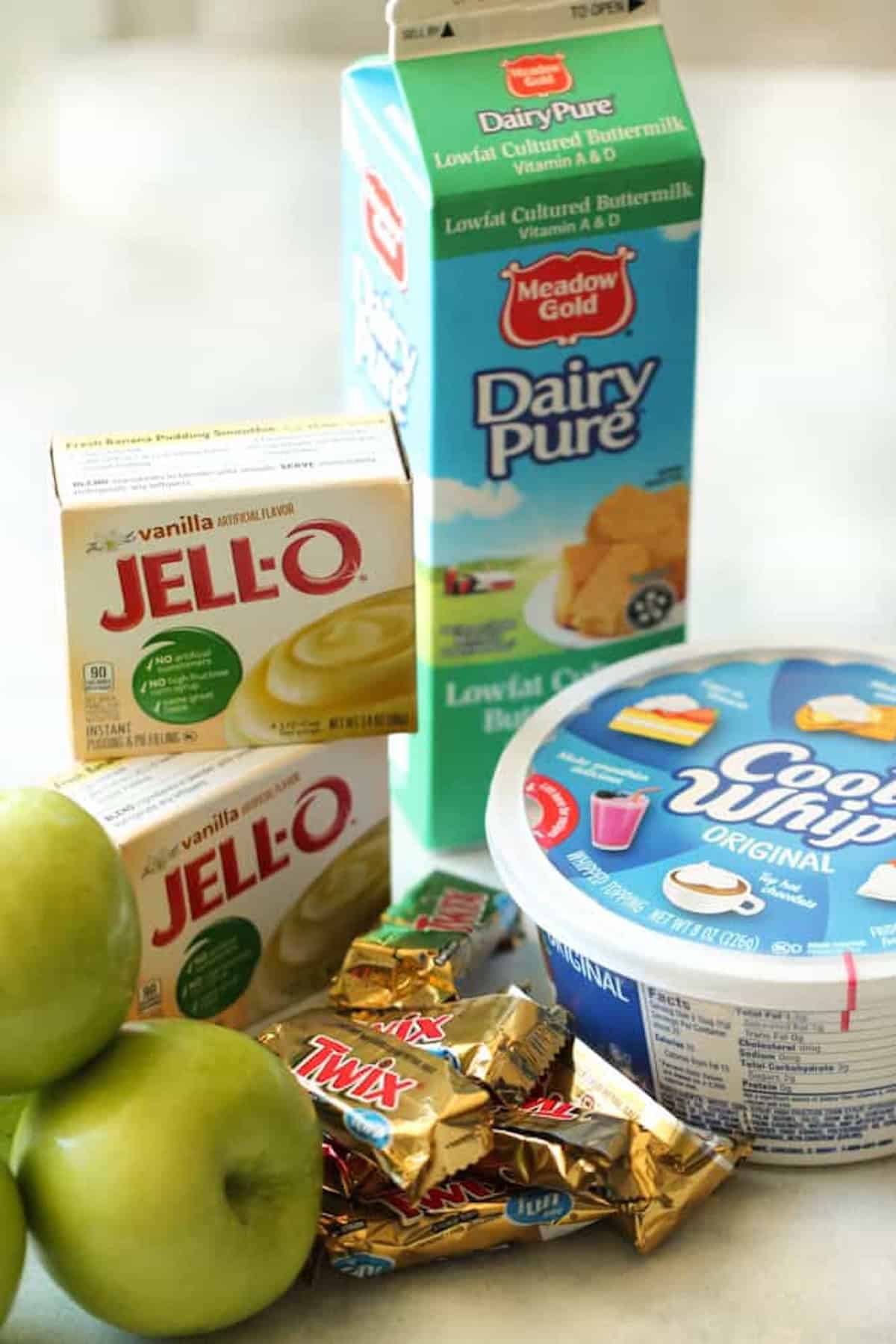 Cartons of buttermilk, boxes of vanilla Jell-O pudding, a tub of Cool Whip, green apples, and wrapped Twix bars are arranged on a counter.