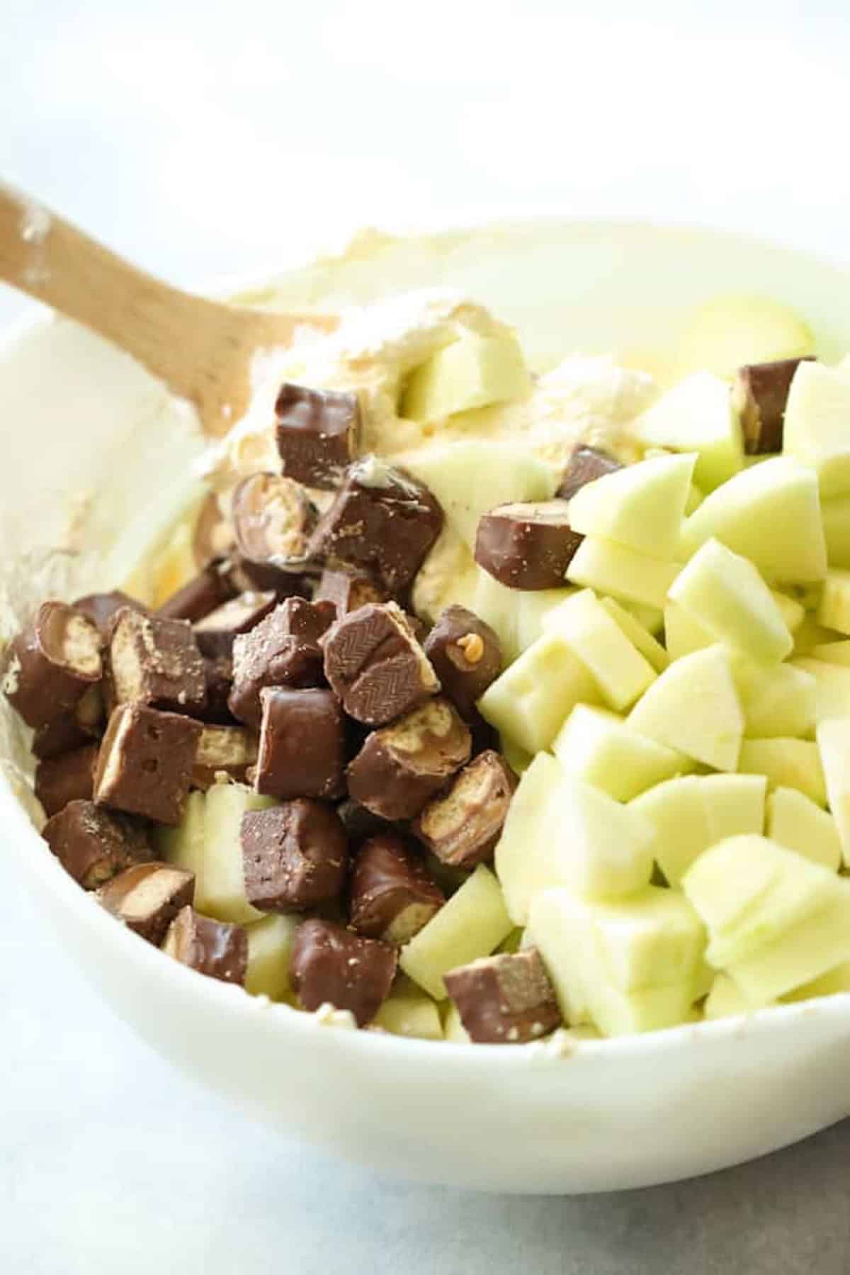 A wooden spoon mixing pudding, cubed apples and chopped mini twix bars together in a mixing bowl.