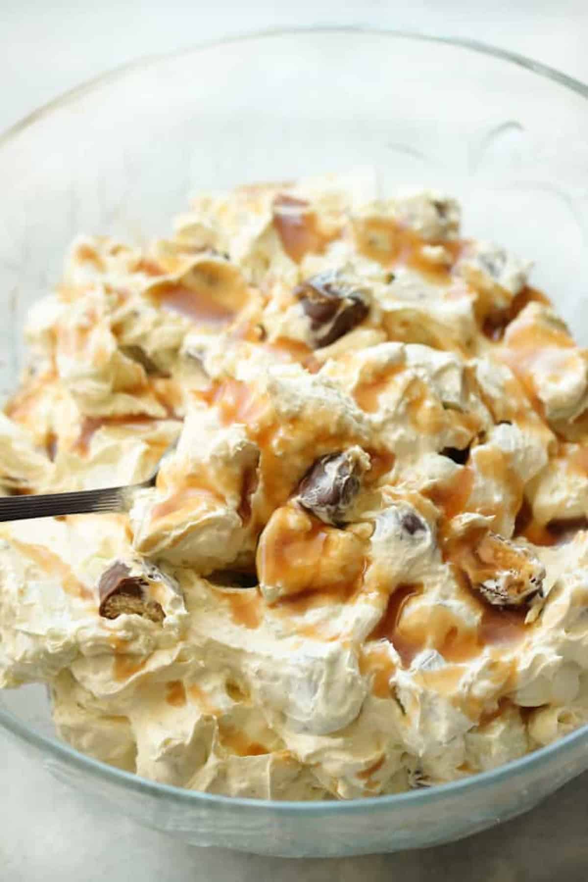 A glass bowl filled with creamy dessert topped with caramel sauce and pieces of dates, with a spoon partially visible on the left side.