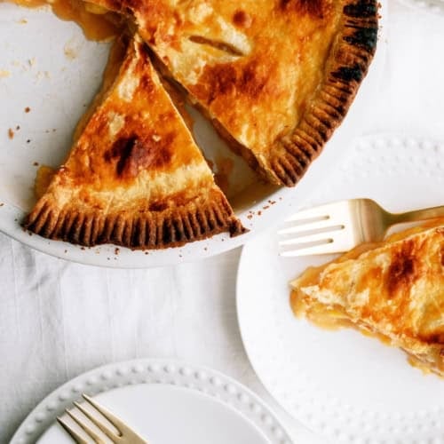 A golden-brown pie with a slice removed sits on a plate; a slice of the pie is served on a white plate beside gold forks on a white tablecloth.