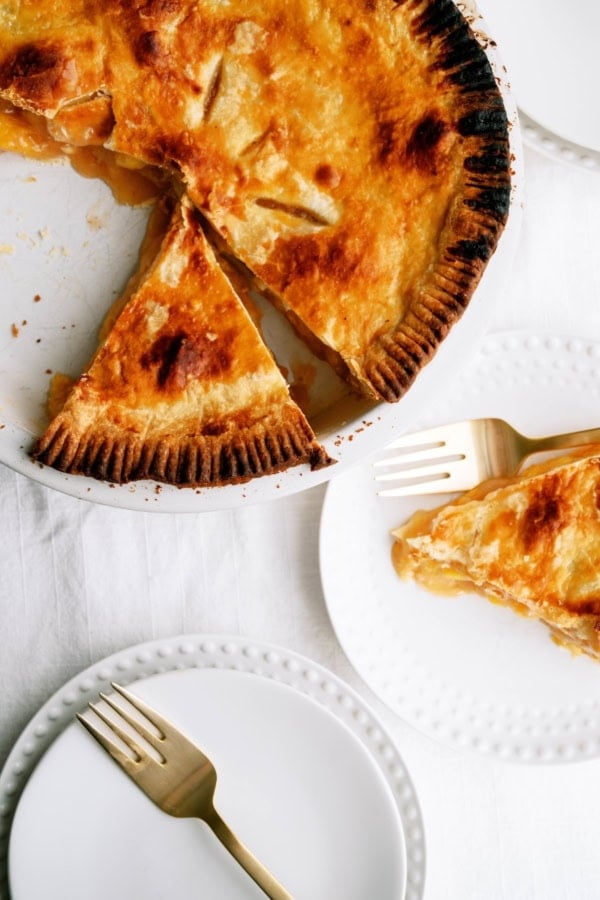 A golden-brown pie with a slice removed sits on a plate; a slice of the pie is served on a white plate beside gold forks on a white tablecloth.