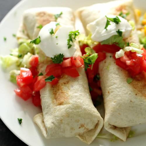 Two burritos topped with chopped tomatoes, sour cream, and cilantro are served on a white plate with lettuce.