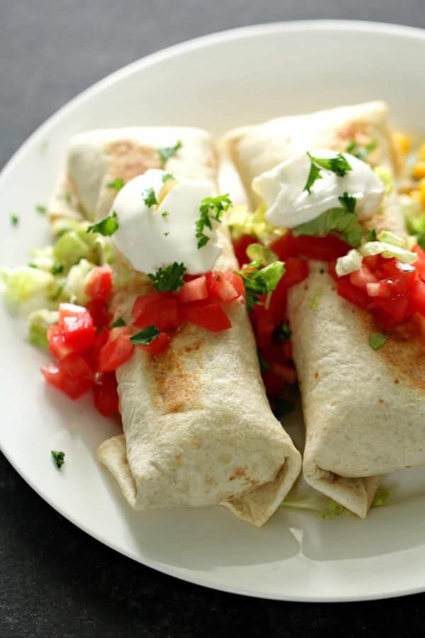 Two burritos topped with chopped tomatoes, sour cream, and cilantro are served on a white plate with lettuce.