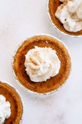 A small pumpkin pie topped with a swirl of whipped cream on a light background.