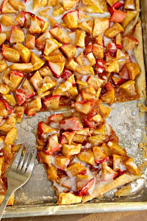 A piece of fruit tart with a combination of sliced apples and strawberries on a flaky crust and drizzled with icing on a baking sheet. A fork is placed beside the tart.