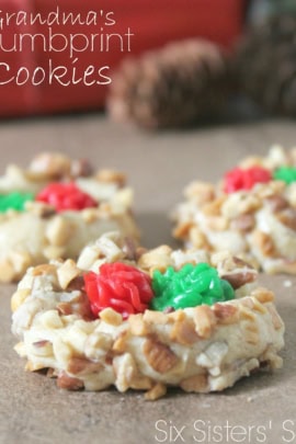 Three thumbprint cookies topped with green and red icing are displayed on a brown surface with pine cones in the background.