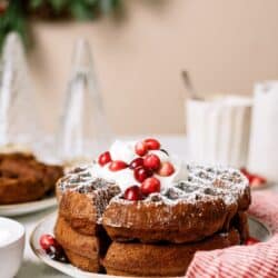 A stack of waffles topped with whipped cream and cranberries on a white plate, with festive decorations and folded red-striped napkin in the background.